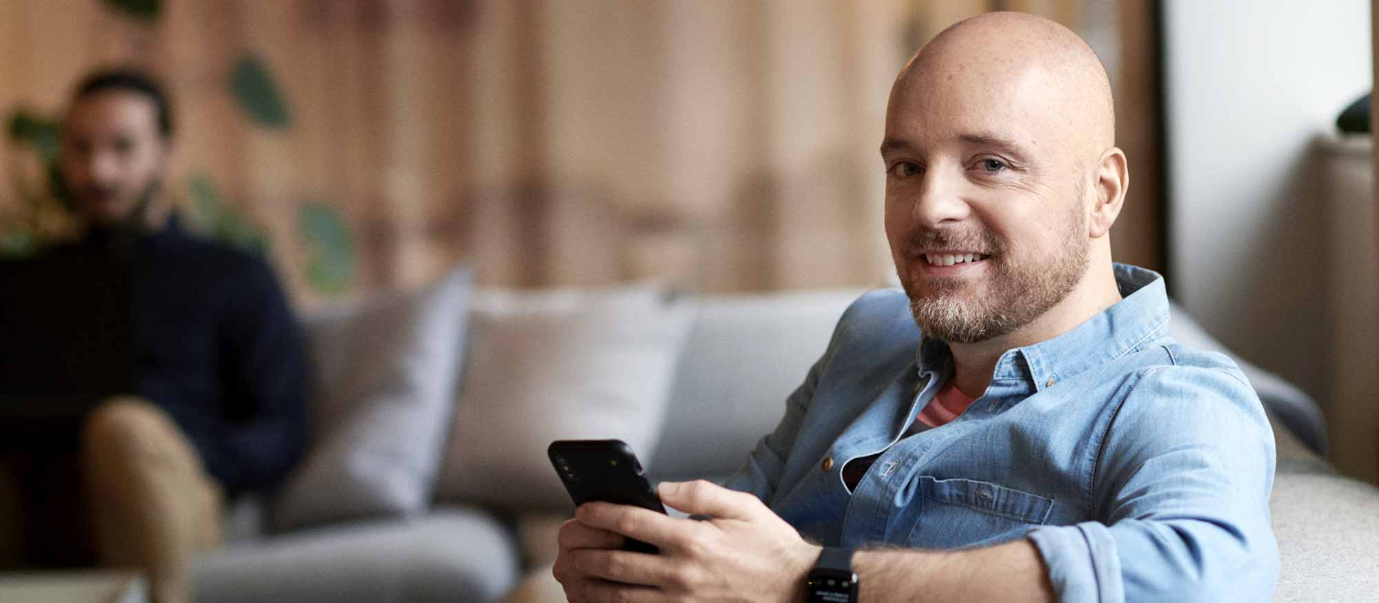 Leende man i jeansskjorta sitter i en grå soffa och håller i sin mobiltelefon
