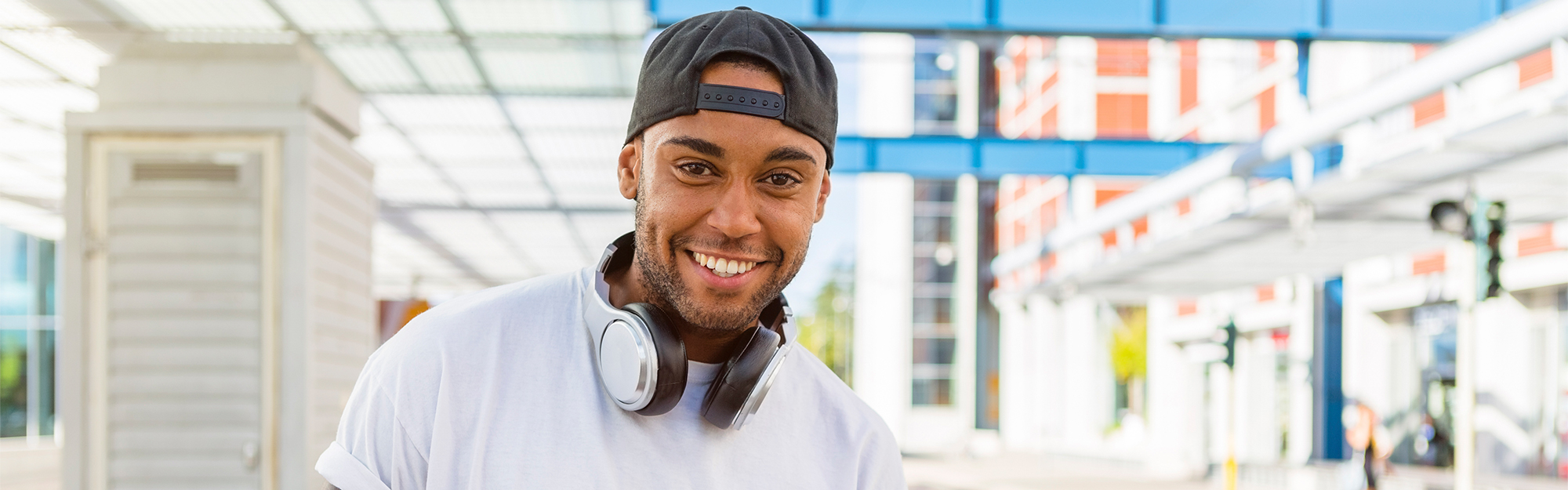 Smiling young man with headphones and smartphone waiting at tram stop.